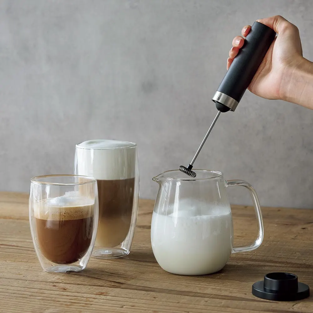  “Mano sosteniendo un espumador de leche sobre una jarra de leche, preparando café con leche espumada”.